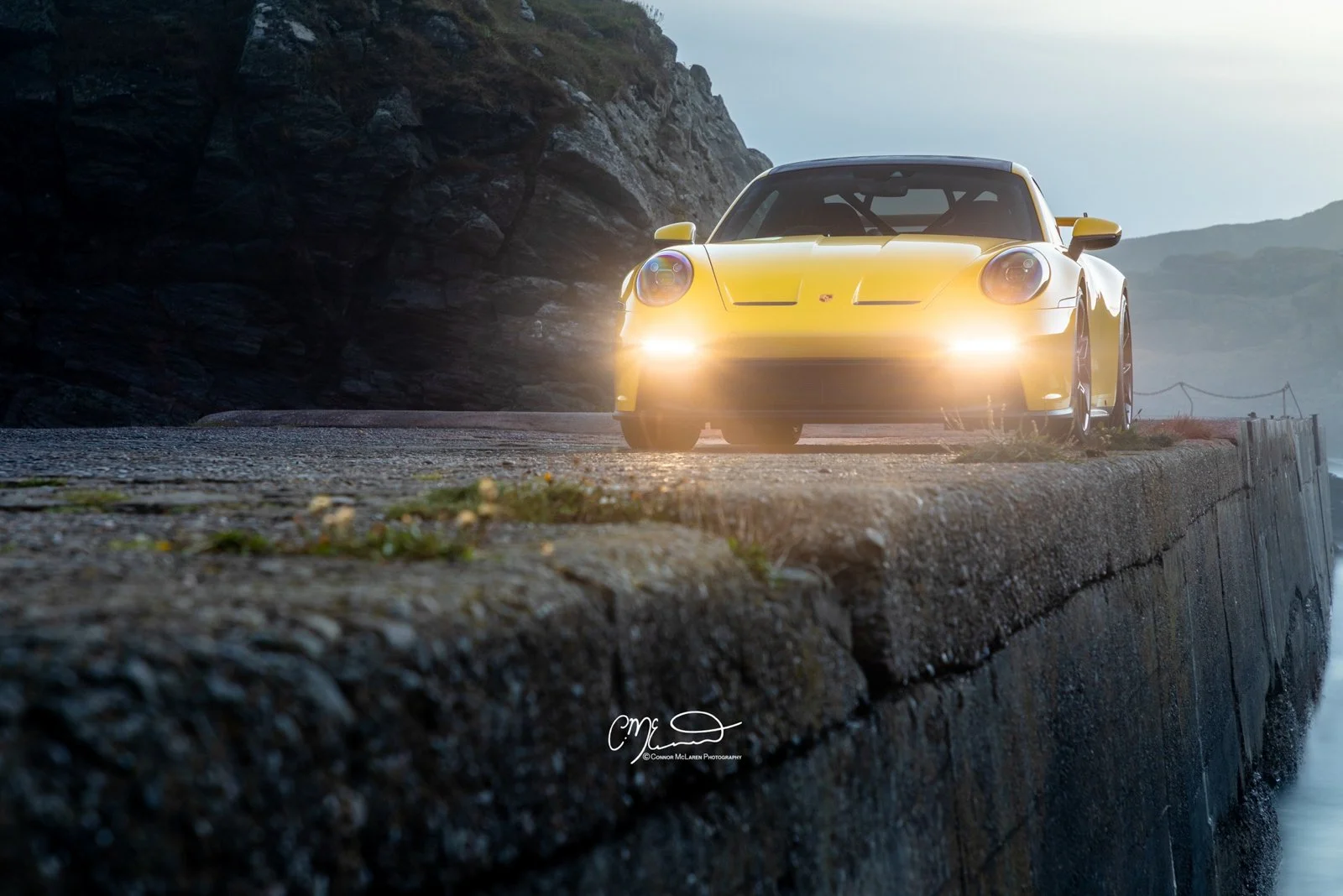 A yellow Porsche GT3 sports car with its headlights on, driving along a narrow, rocky road by a body of water with mountains in the background.