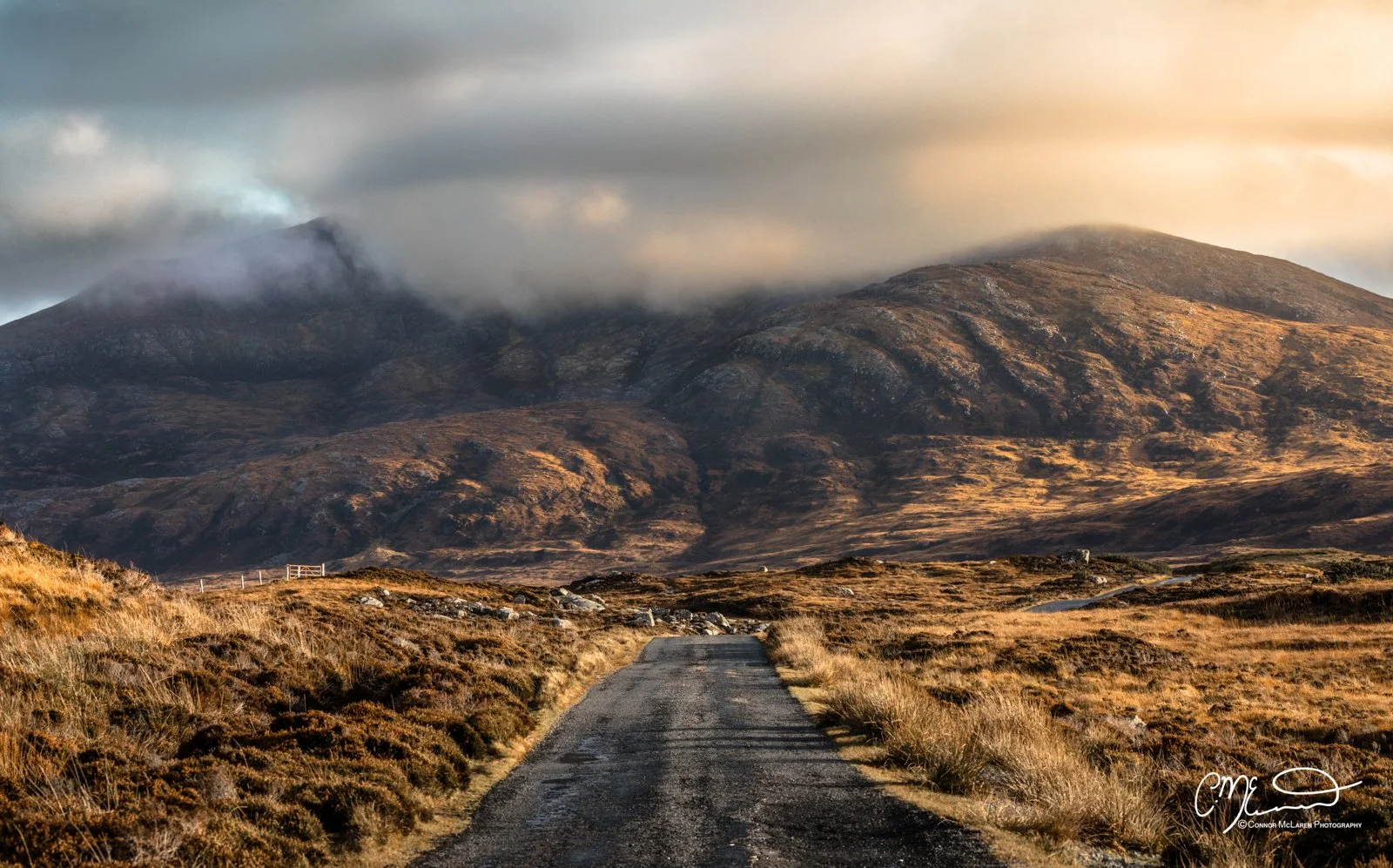 Scotland, Lewis Harris & Outer Hebrides, South Uist. A rural gravel road leading towards mountains with cloudy sky and rolling hills, in a landscape with dry grass and shrubs.