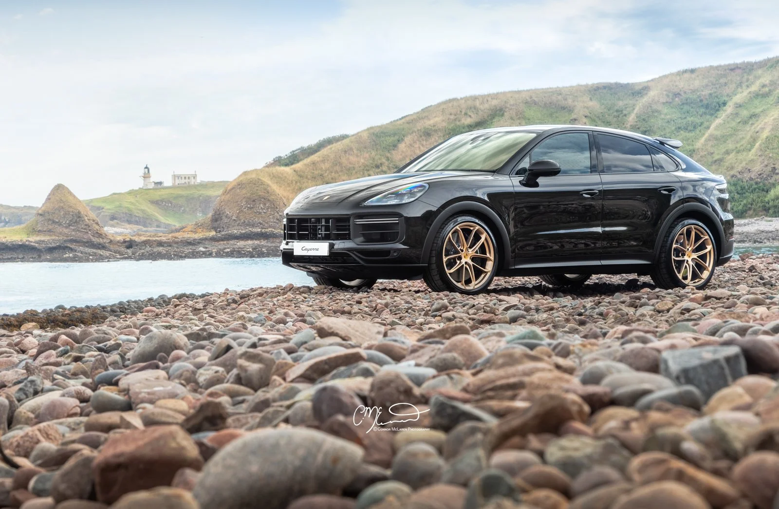 A black Porsche Cayenne Turbo GT with gold wheels parked on a rocky beach near the water, with green hills and a lighthouse in the background.