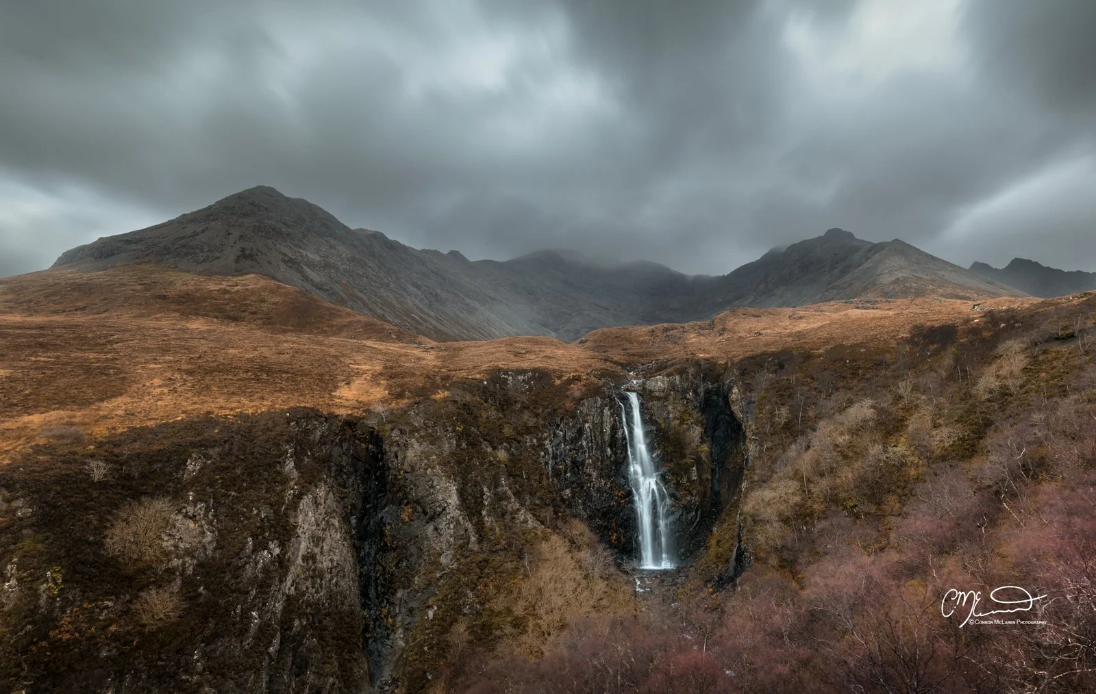 Scotland, Isle of Skye, Fairy Pools. A scenic mountain landscape with dark clouds, brown hills, and a waterfall cascading down a rocky cliff.
