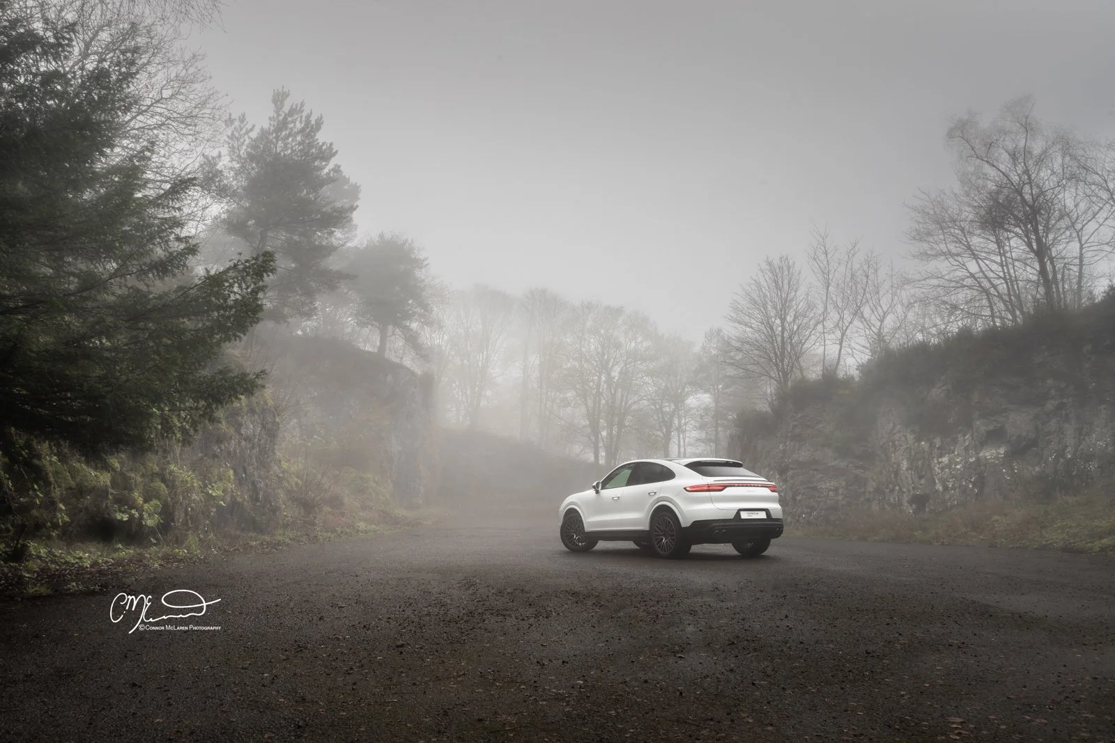 A white Porsche Cayenne coupe parked on a foggy gravel road surrounded by bare and evergreen trees and rocky hills.