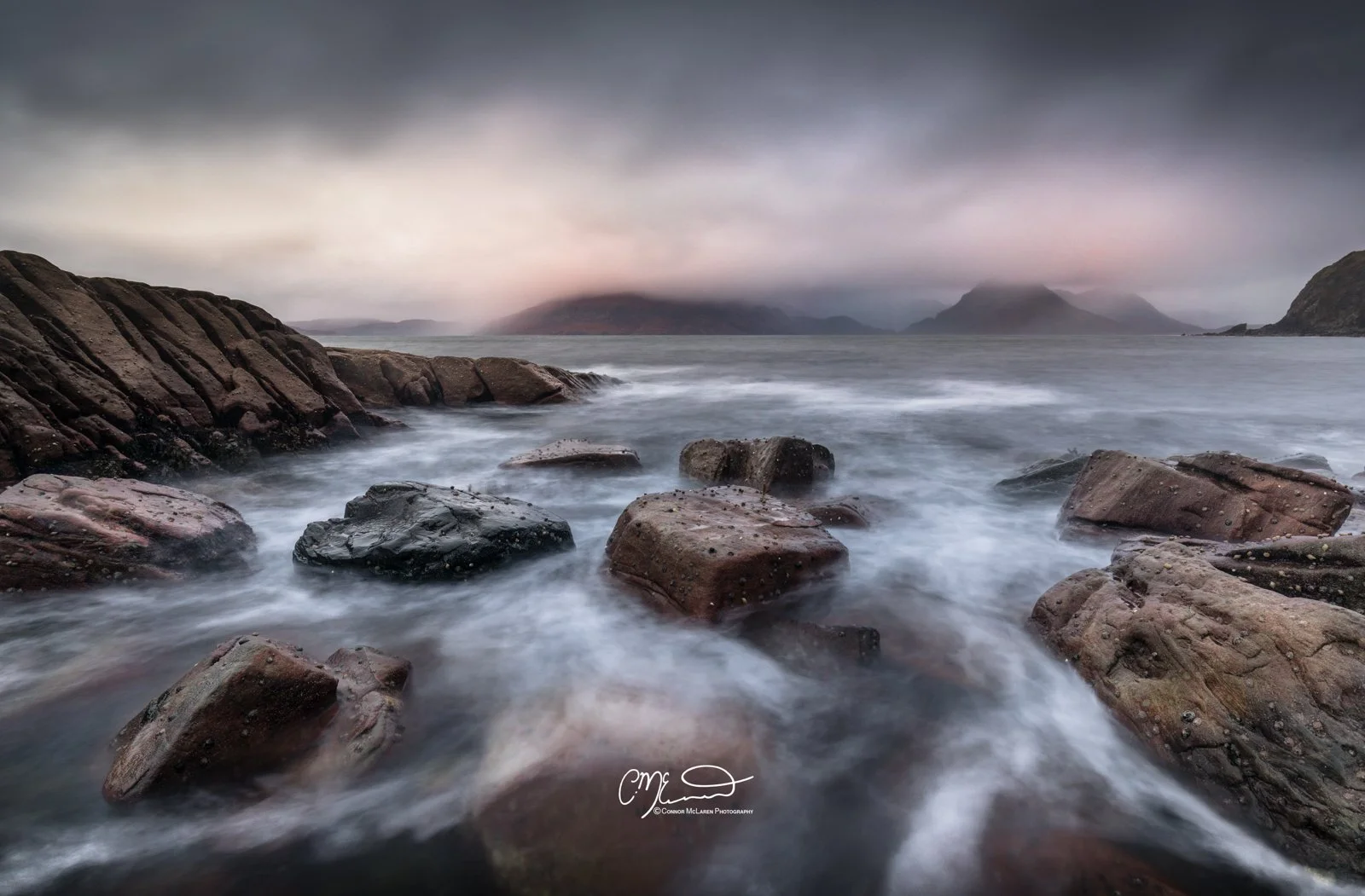 Scotland, Isle of Skye, Elgol. Rocky shoreline with water washing over rocks under a cloudy sky, mountains in the distance.