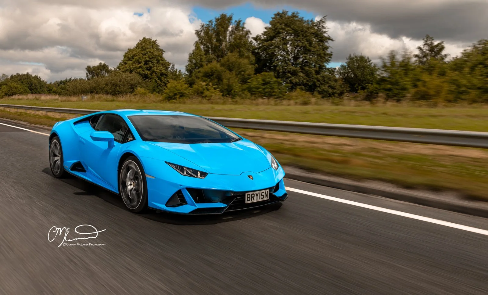 A blue Lamborghini huracan evo sports car driving on a highway with a grassy embankment and trees in the background, under a partly cloudy sky.