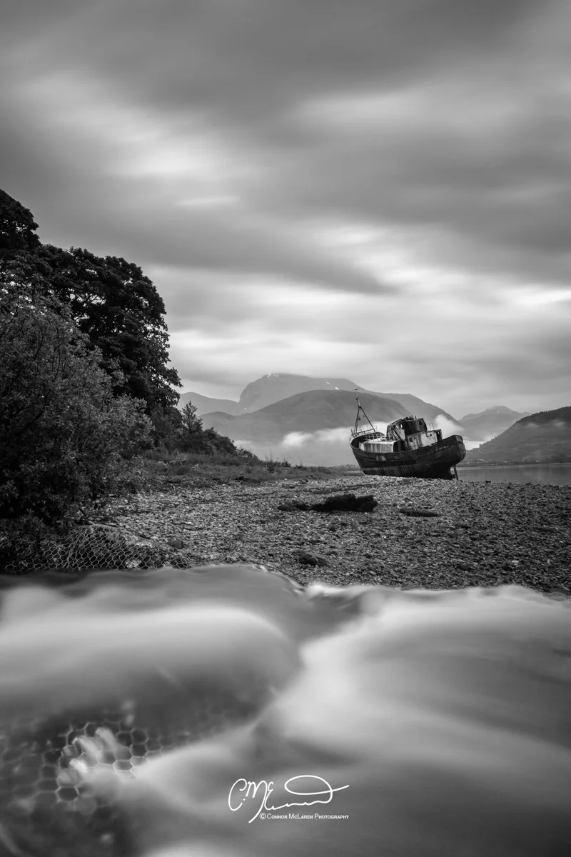 Scotland, Fort William, Ben Nevis. A black and white photo of a coastal landscape with an abandoned boat on the rocky shoreline, mountains in the background, and cloudy sky above.