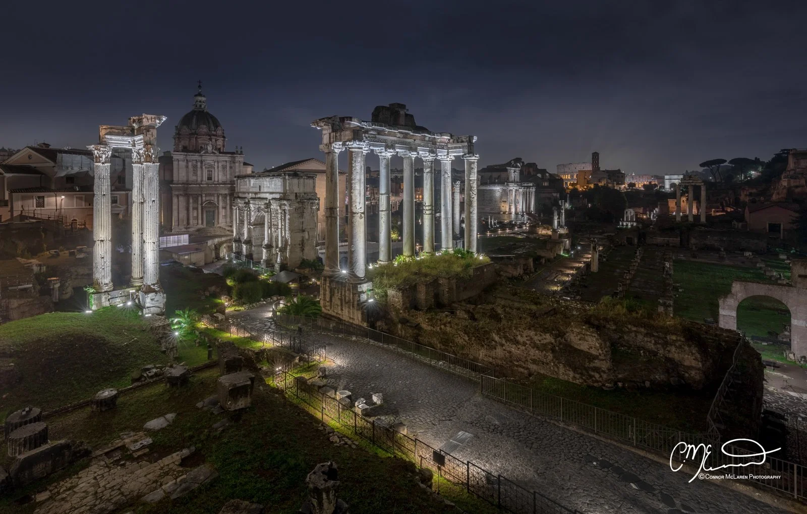 Roman Forum, Rome & the vatican. Ancient Roman ruins at night illuminated by lights, with city buildings in the background and a dark sky.