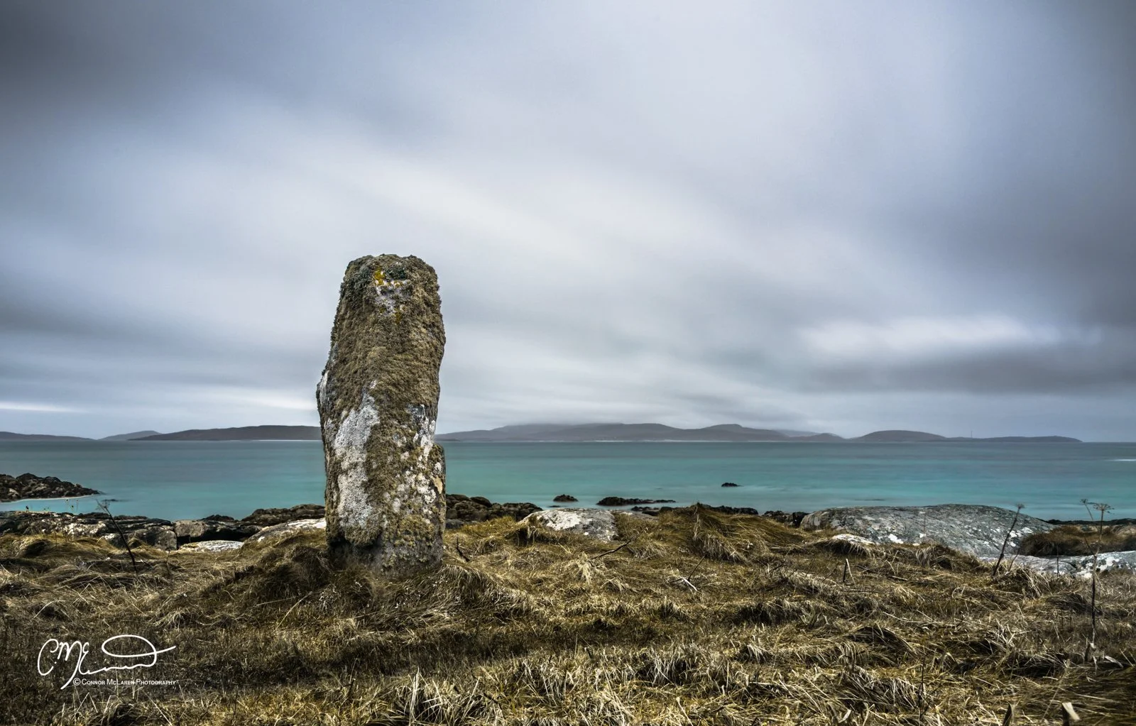 Scotland, Lewis Harris & Outer Hebrides, South Uist. A large moss-covered standing stone on a grassy shoreline with an ocean and distant islands under a cloudy sky.