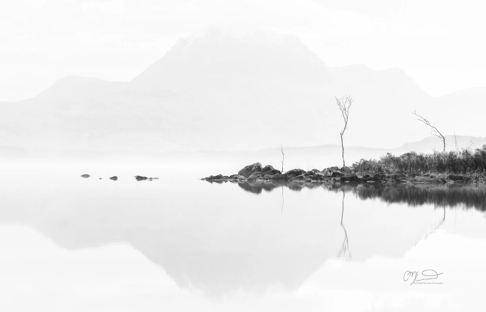 Scotland, Loch Maree. Black and white landscape photo of a calm lake with rocks and leafless trees along the shoreline, mountain in the background, reflected in the water.
