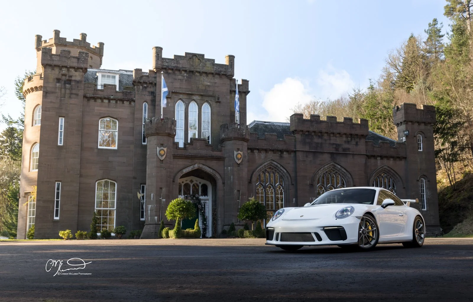 A white Porsche 911 GT3 sports car parked in front of a large, castle-like stone building with towers, arched windows, and flags, surrounded by trees.