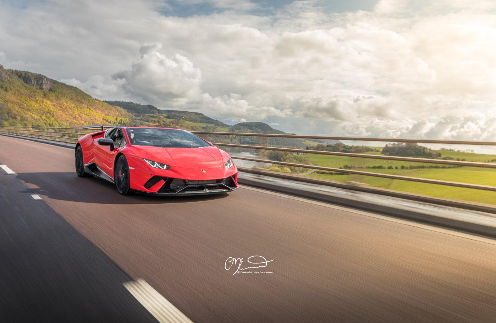 Lamborghini Performante Spyder. Red Lamborghini sports car driving on a highway with green hills and cloudy sky in the background.