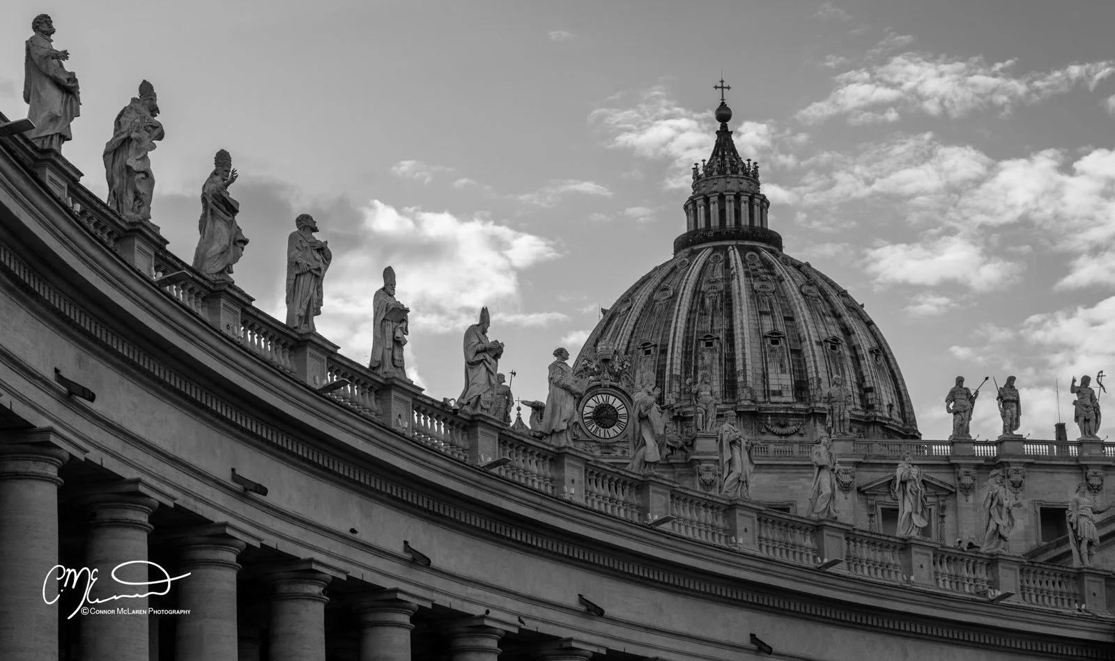 Roman Forum and colosseum, Rome & the vatican. Black and white photograph of St. Peter's Basilica with a series of statues along the edge of the roof, a clock, and a large dome with a cross on top, under a partly cloudy sky.