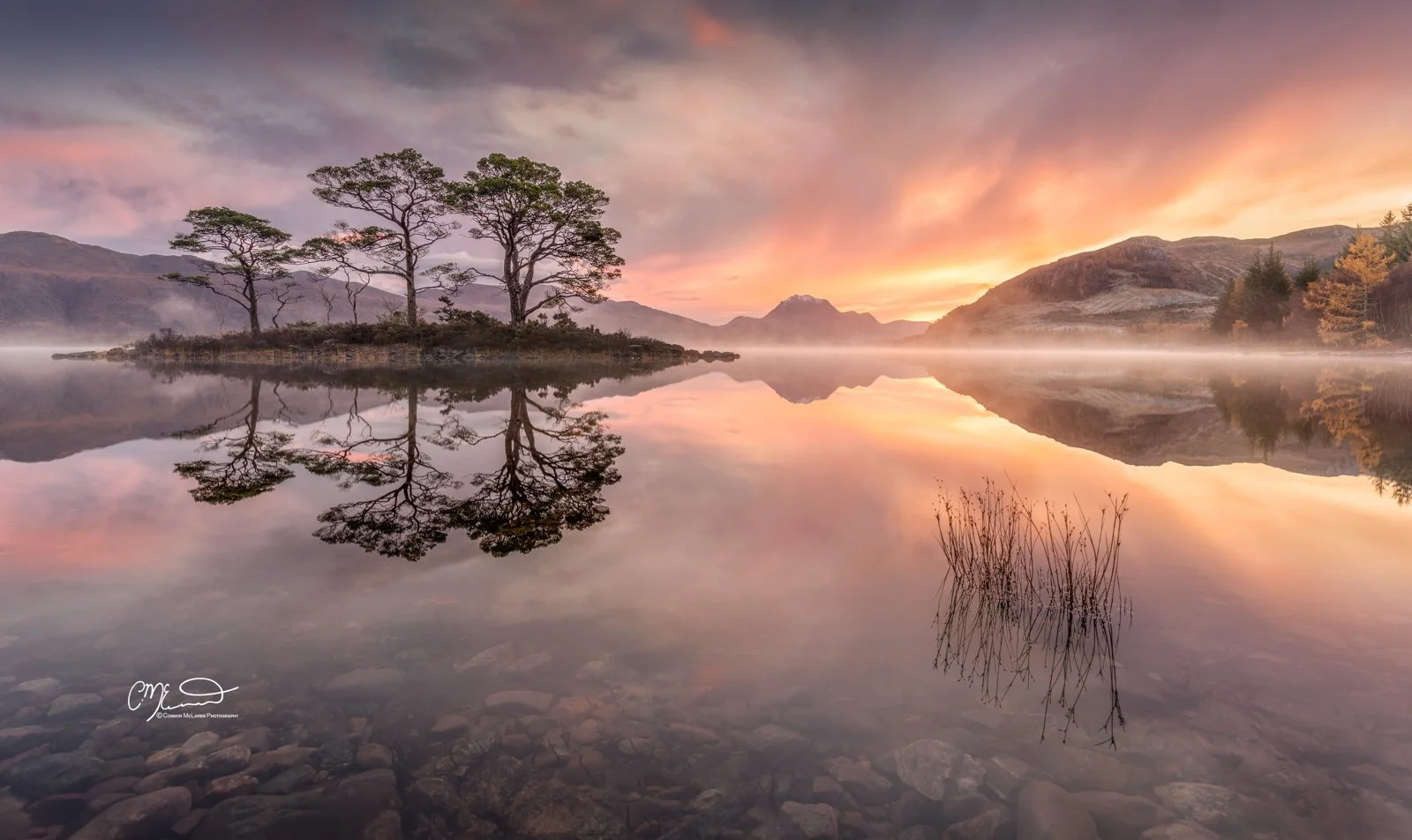 Scotland, north coast 500, NC500, Loch Maree. A calm lake at sunrise with mountains in the background. Trees and their reflections are visible in the water, with mist rising from the lake's surface.