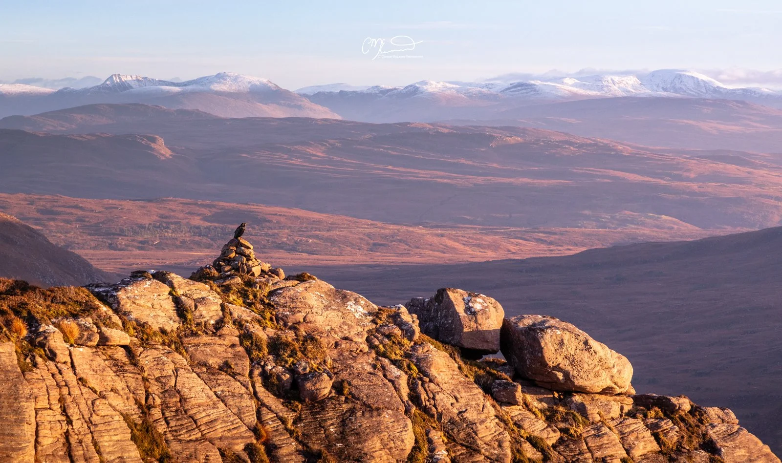 Scotland, north coast 500, NC500, Assynt. Stac Pollaidh. Scenic view of rocky mountain foreground with a cairn, vast valley below, distant mountains with snow caps, and a clear sky.