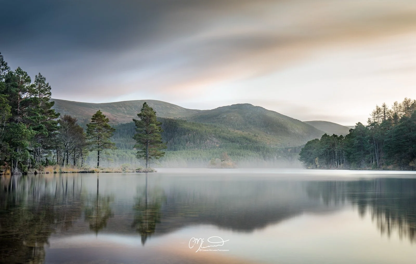 Scottish Central Highlands, Scotland, Cairngorms Loch an Eilean. A serene lake reflecting a line of pine trees and forested hills in the distance under a cloudy sky.