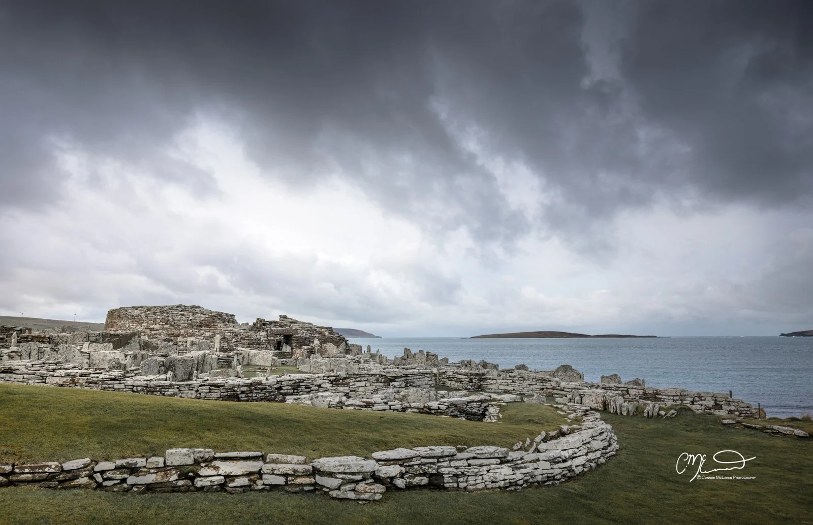 Broch of Gurness, Orkney, Scotland. Ancient stone ruins on a grassy coastline with a cloudy sky and the ocean in the background.