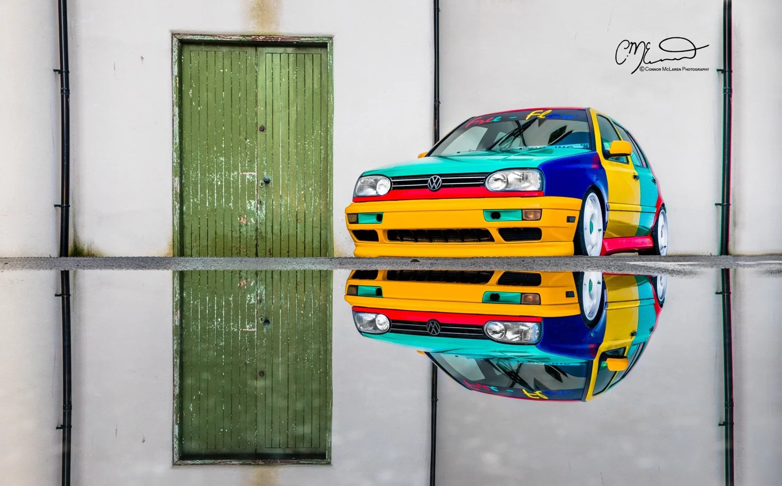 Colorful Volkswagen Golf Harlequin car with a vibrant, rainbow-like paint job parked in front of a white wall with a green door, and its reflection is captured in a puddle on the ground.