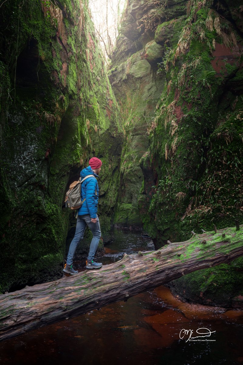 Scottish Lowlands, Scotland, devils pulpit, Outlander filming location. A person wearing a red hat and blue jacket standing on a fallen log across a small stream in a moss-covered canyon with steep rocky walls and greenery.