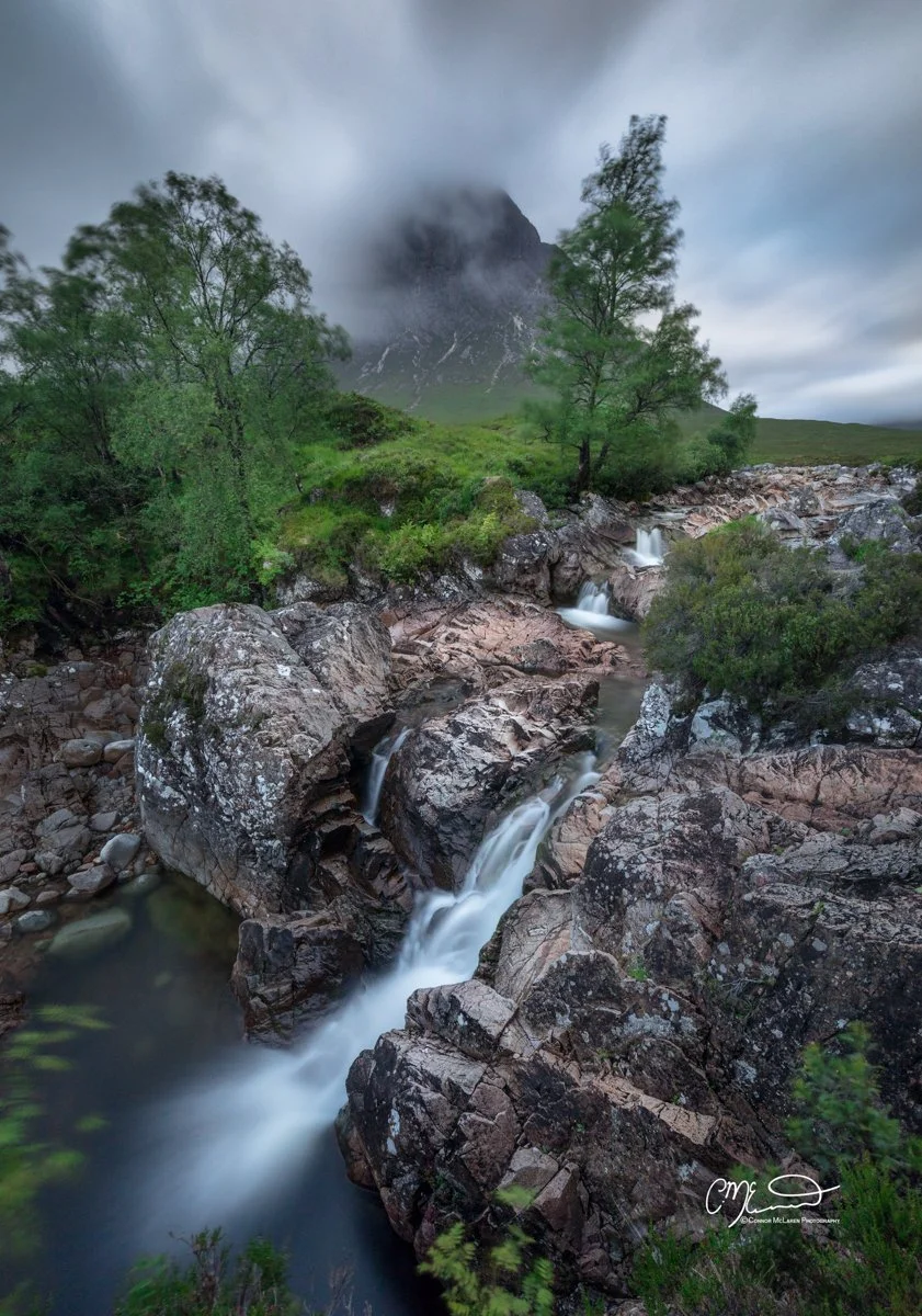 Glen Coe and Glen Etive, Western Highlands, Scotland. A scenic landscape featuring a mountain shrouded in clouds, tall green trees, and a flowing creek cascading over rocks in a lush, green wilderness.