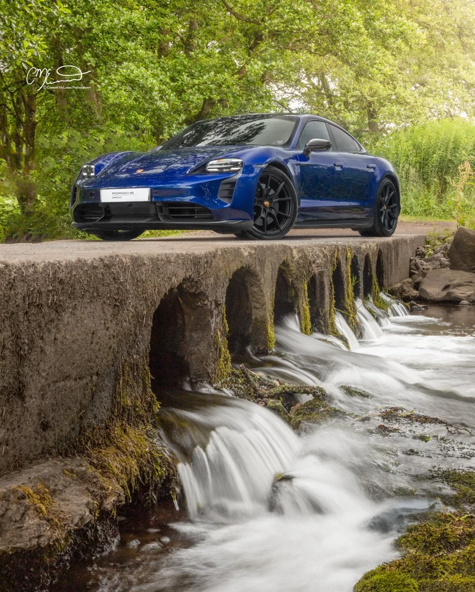 A blue Porsche Taycan GTS sports car parked on a concrete bridge over a flowing small stream, surrounded by green trees and foliage.