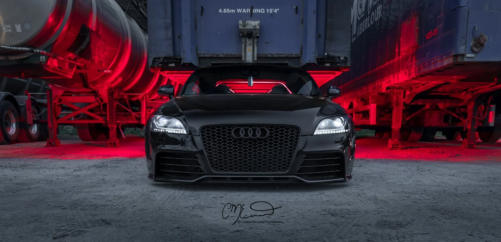 Custom Audi TTRS in black, bagged air suspension. A black Audi sports car parked on a gravel lot with red LED lighting on the background, flanked by two large trailers.
