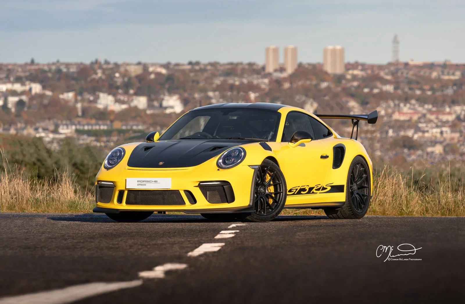 A yellow and black Porsche 911 GT3 RS sports car parked on a road with a cityscape in the background.