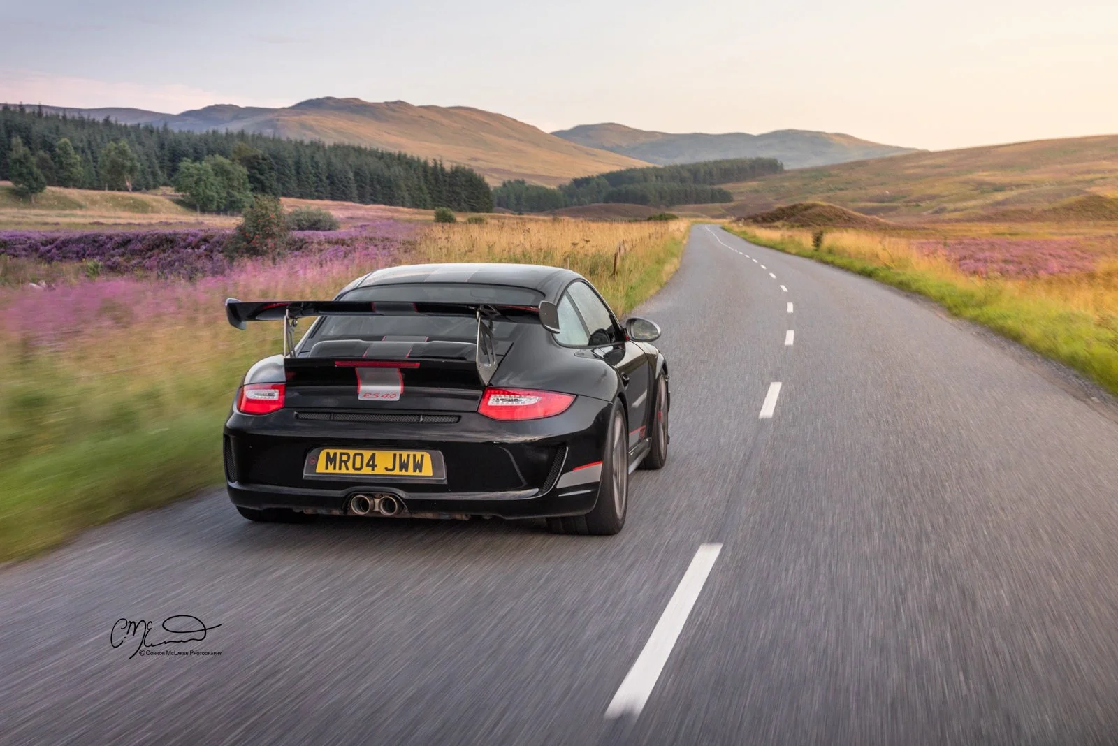 Porsche 911 GT3 RS 4.0L. A black sports car with a rear spoiler driving on a rural road through scenic countryside with hills and purple flowers