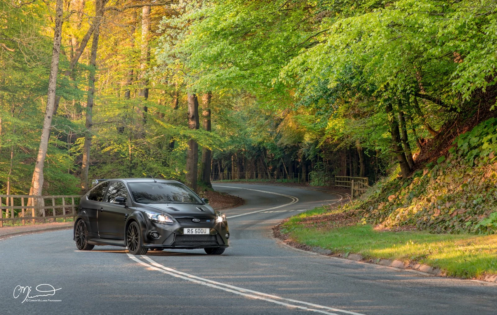 A black Ford Focus RS500 is driving on a winding forest road surrounded by trees with green and yellow leaves.