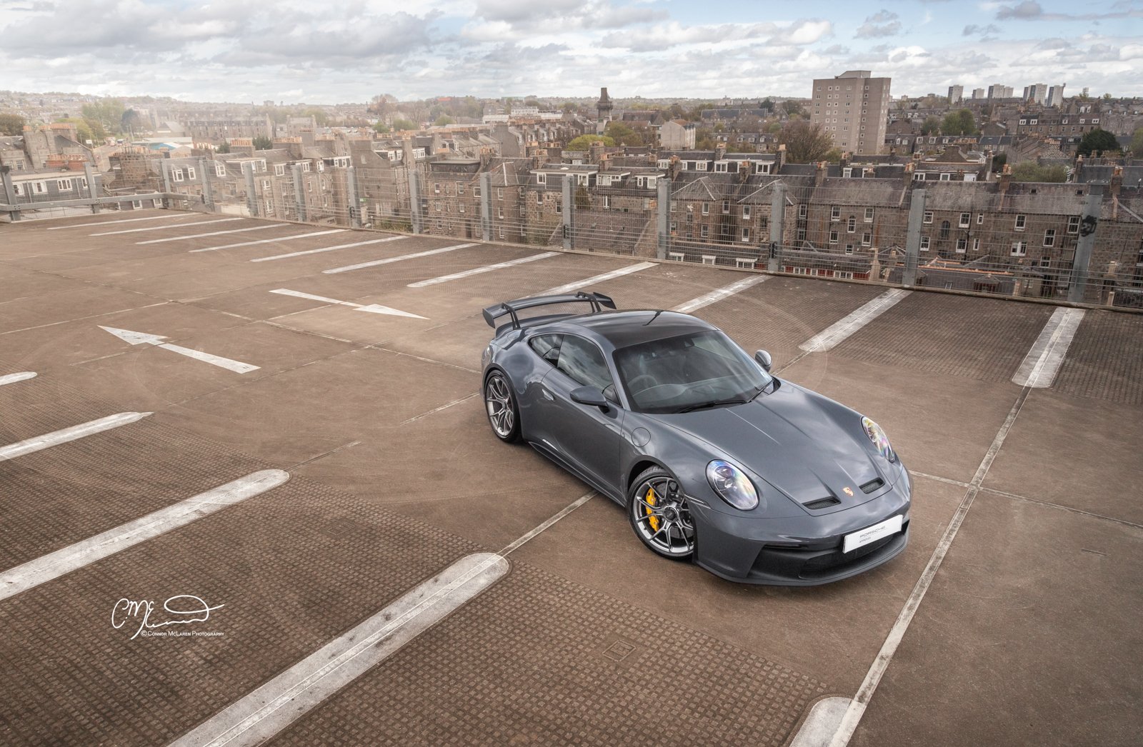 A gray Porsche 911 GT3 sports car parked in an empty rooftop parking lot with city buildings and cloudy sky in the background.