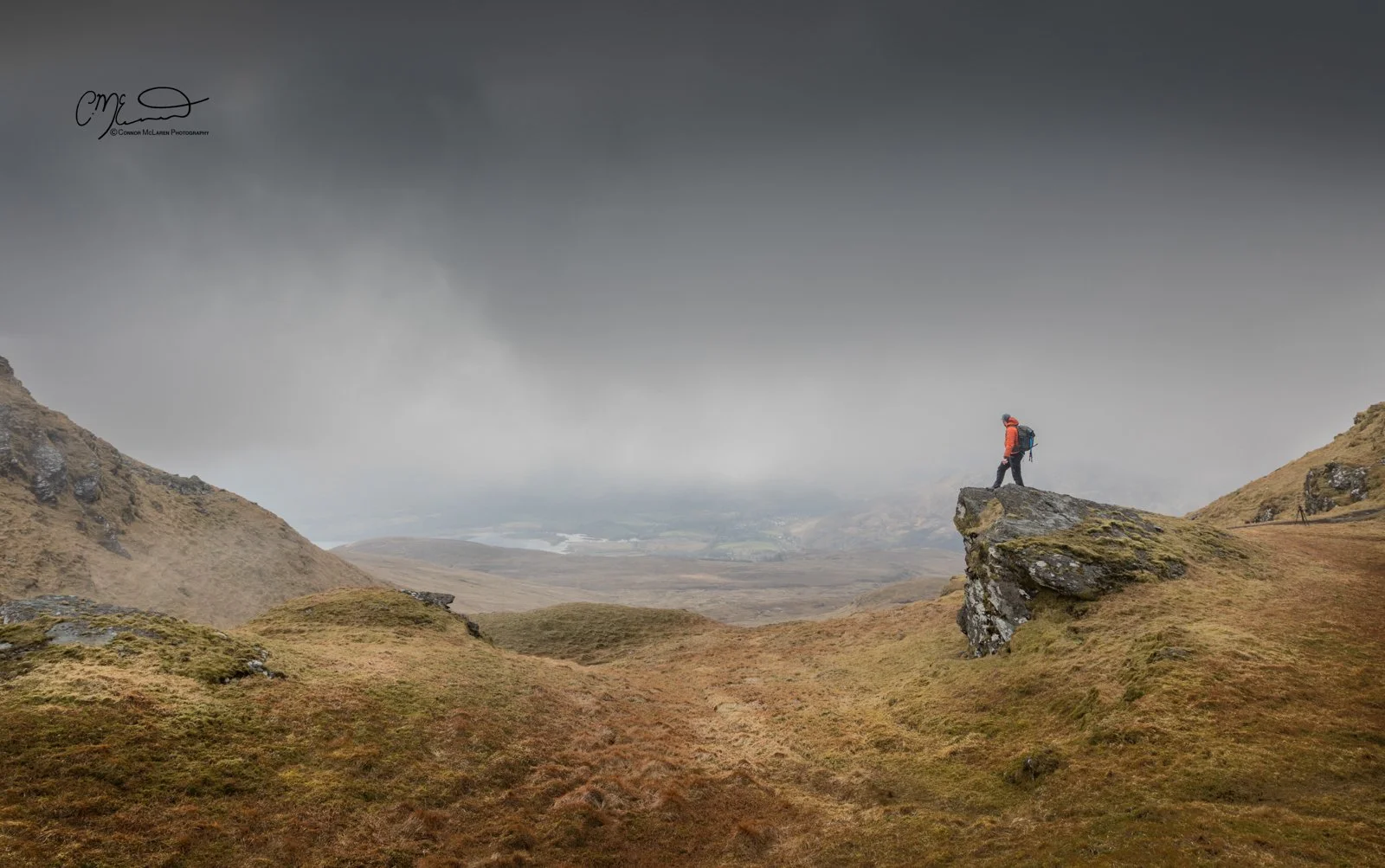 Scottish Central Highlands, Scotland, Perthshire munro. A person with a red jacket and backpack standing on a rocky ledge in a mountainous landscape under a cloudy sky.