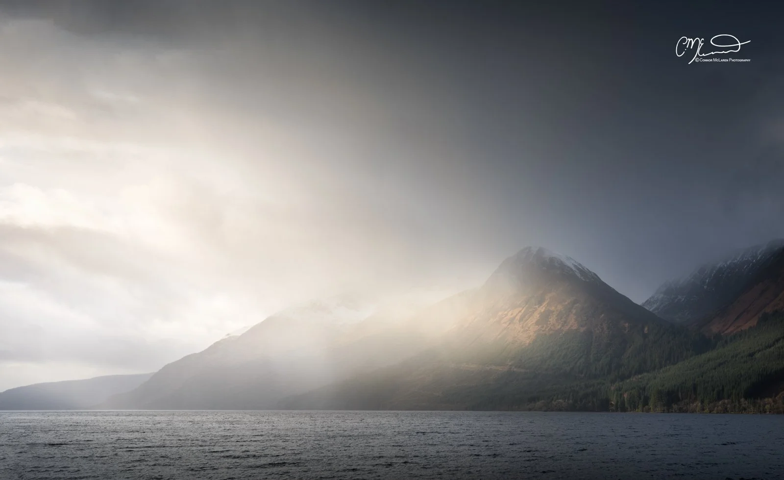 Loch, Western Highlands, Scotland. A vast body of water with mountains in the background, some snow on the peaks, and a cloudy sky overhead.