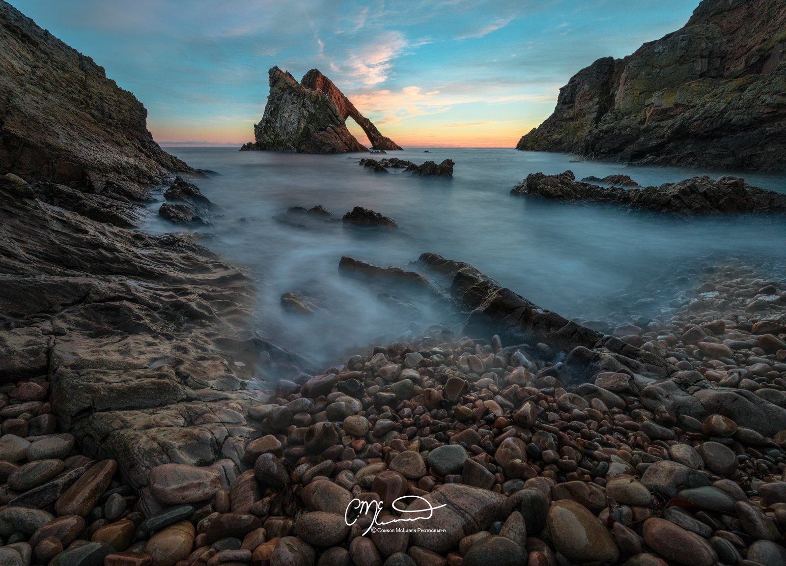 Bow Fiddle Rock, Morrayshire, North East Scotland. Scenic coastal landscape at sunset with large rocks in the foreground, a rocky island in the background, and misty water flowing over the rocks.