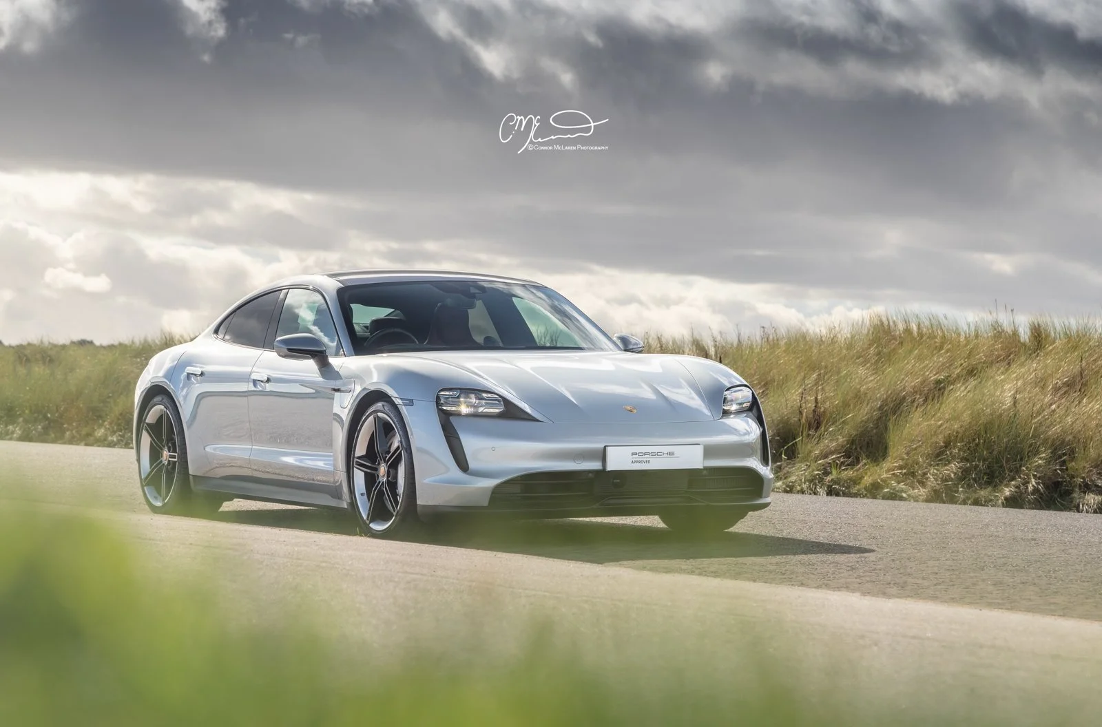 A silver Porsche Taycan sports car on a rural road with grass and cloudy sky in the background.