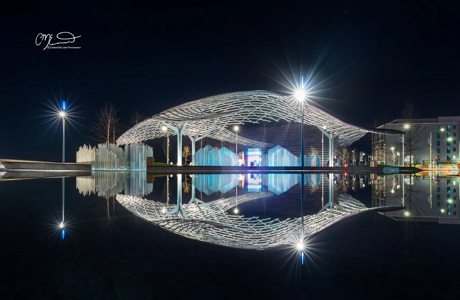Dundee V&A, North East Scotland. Night view of a modern architectural pavilion with a wavy roof structure, illuminated with white lights, reflected in a body of water, with street lamps and trees in the background.