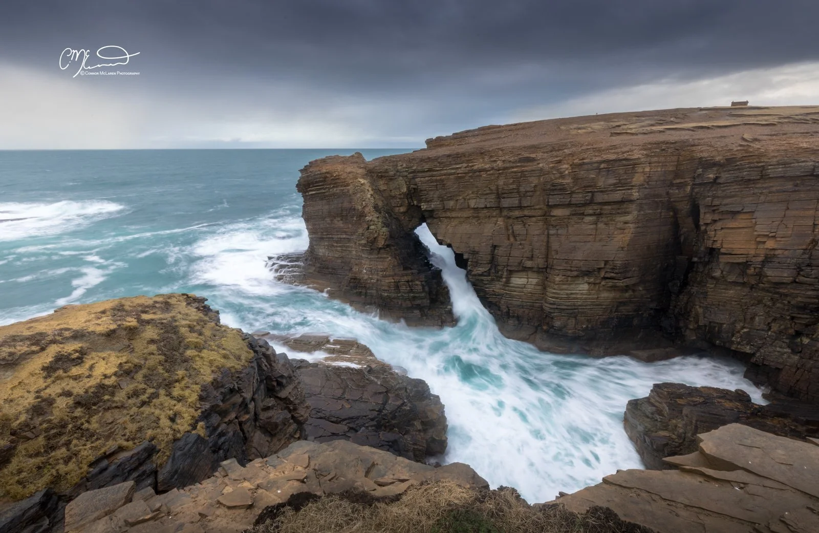 Yesnaby, rainbow, Orkney, Scotland. Coastal scene with waves crashing against layered rock formations under a dark, stormy sky