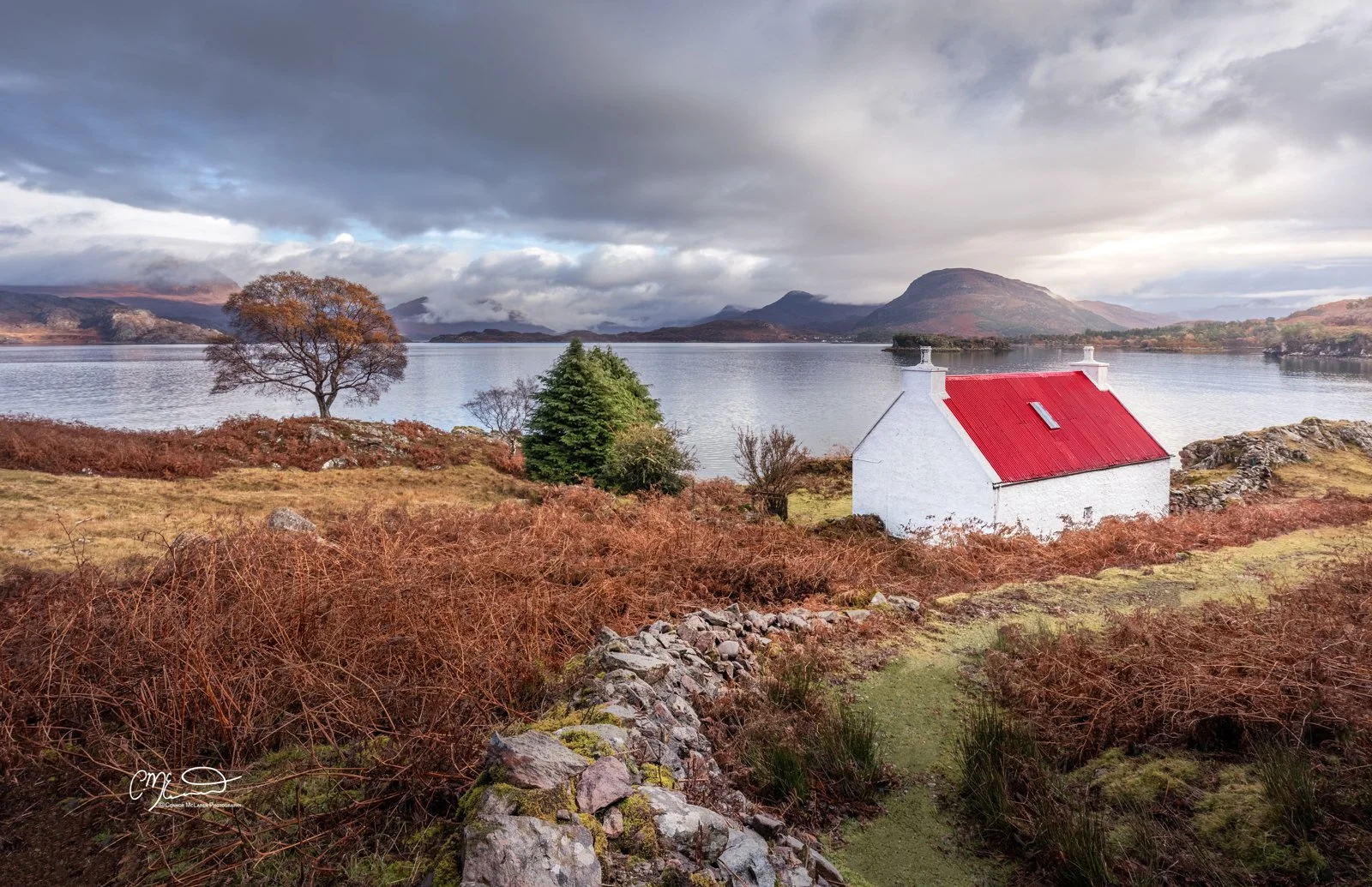 Scotland, north coast 500, NC500, Shieldaig. A small white house with a red roof near a body of water surrounded by autumn-colored shrubs, with mountains and stormy clouds in the background.
