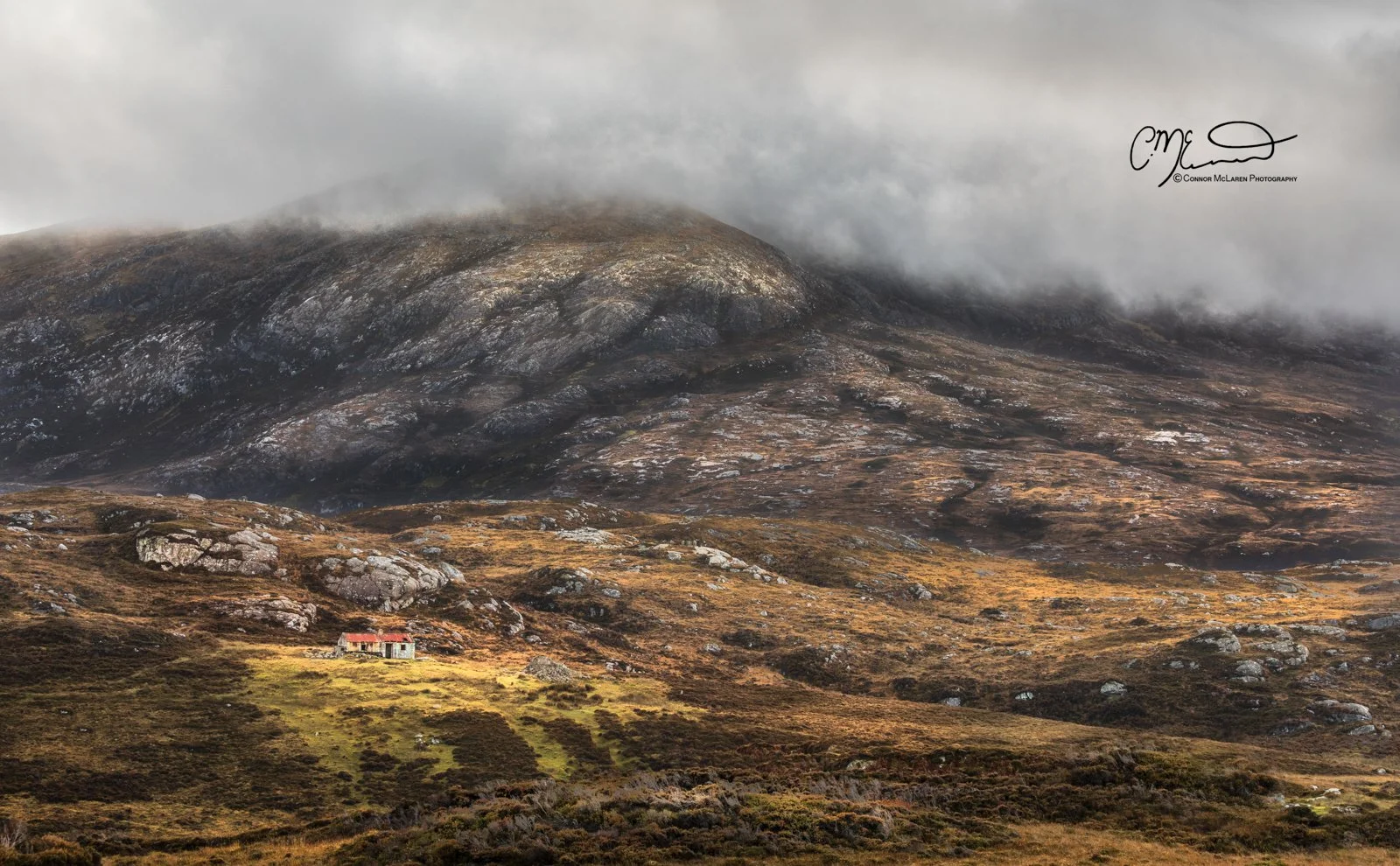 Scotland, Lewis Harris & Outer Hebrides, South Uist. A landscape of rolling hills and mountain with fog hovering over the top, a small house with a red roof nestled among the grassy fields.