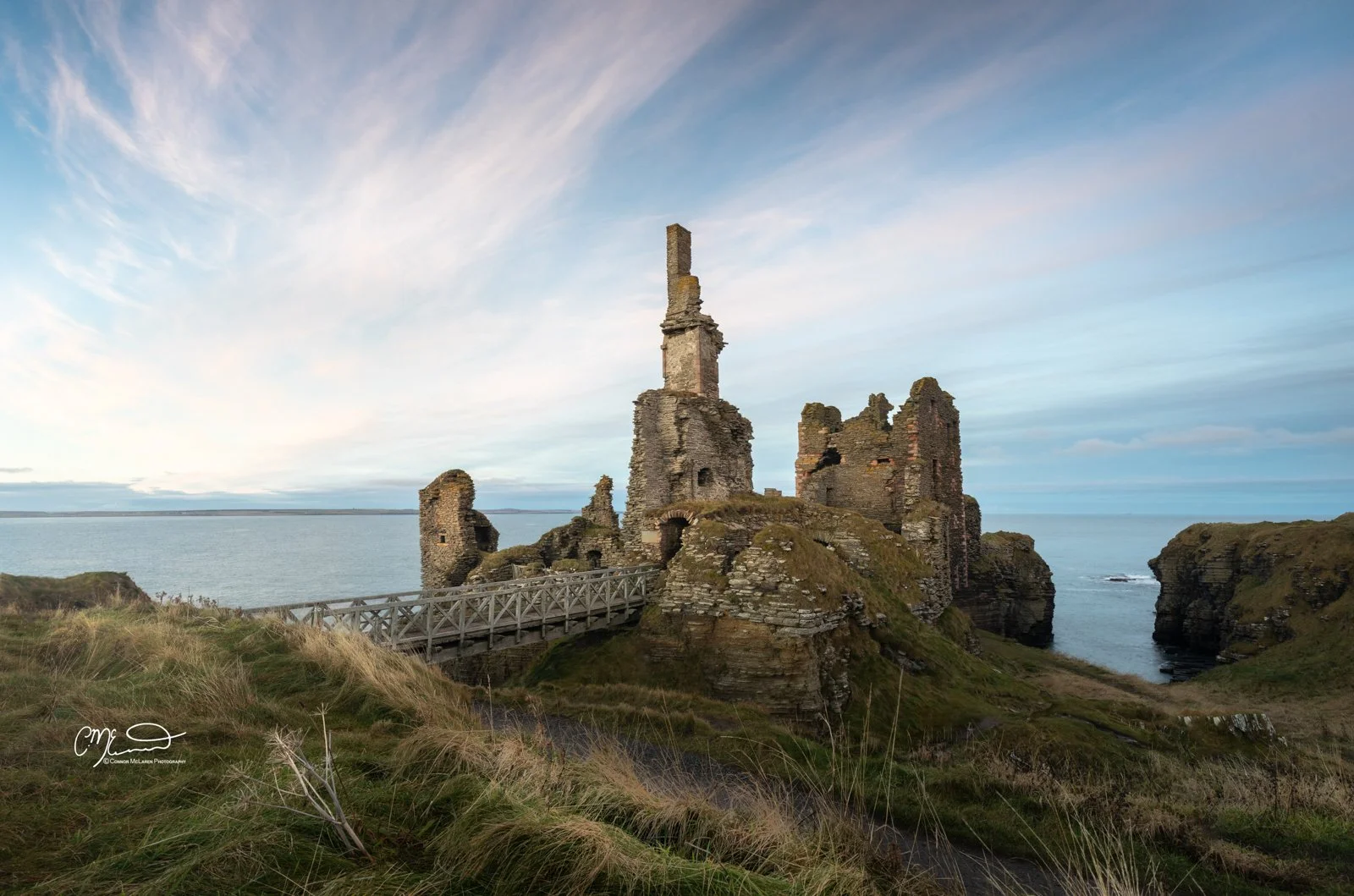 Scotland, north coast 500, NC500, Castle Sinclair Gringoe. Ruins of a castle on a rocky cliff overlooking the ocean under a partly cloudy sky, with a wooden bridge leading to the castle entrance.