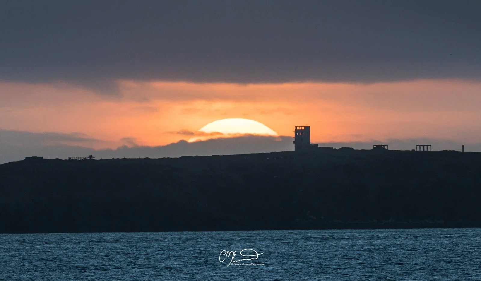 Orkney, Scotland. Sunset over water with silhouetted shoreline and buildings, dark clouds overhead.