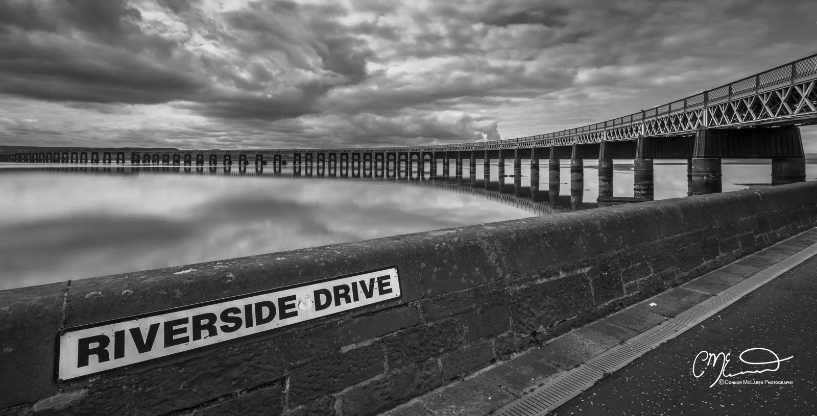 Scotland, Riverside drive Dundee. Black and white photo of a bridge over a river with a cloudy sky. A sign on a stone wall reads "RIVERSIDE DRIVE".