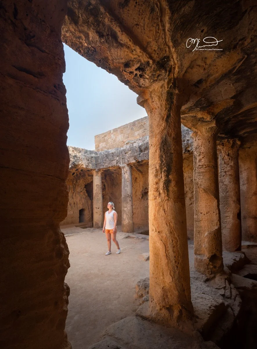 Tomb of the Kings, Cyprus. A woman standing inside the ancient ruins of a stone structure, surrounded by tall, weathered columns with a partially open sky above.