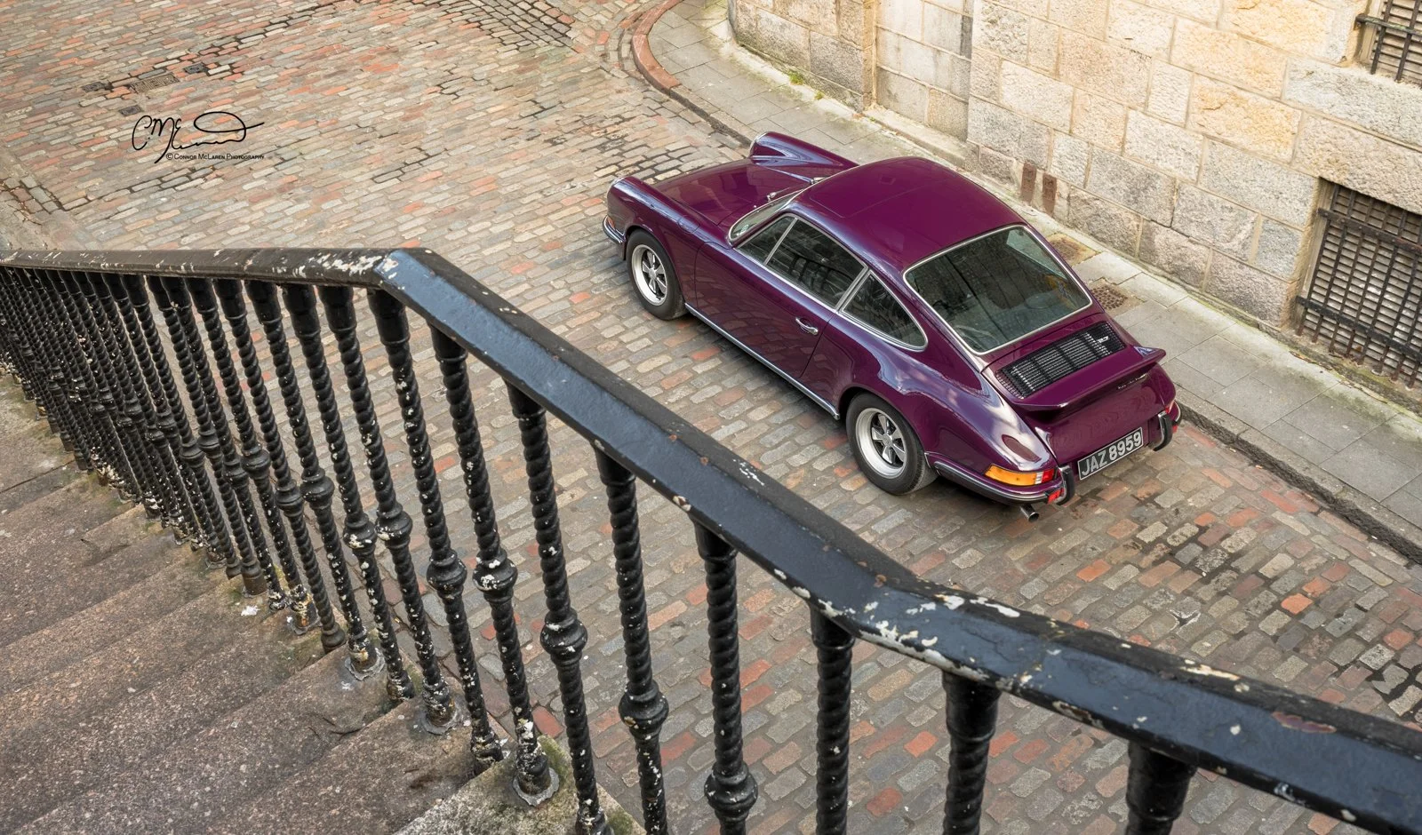 Porsche 911, 2.7RS. A vintage purple Porsche car parked on a cobblestone street viewed from above, with a black iron railing and stone building in the background.