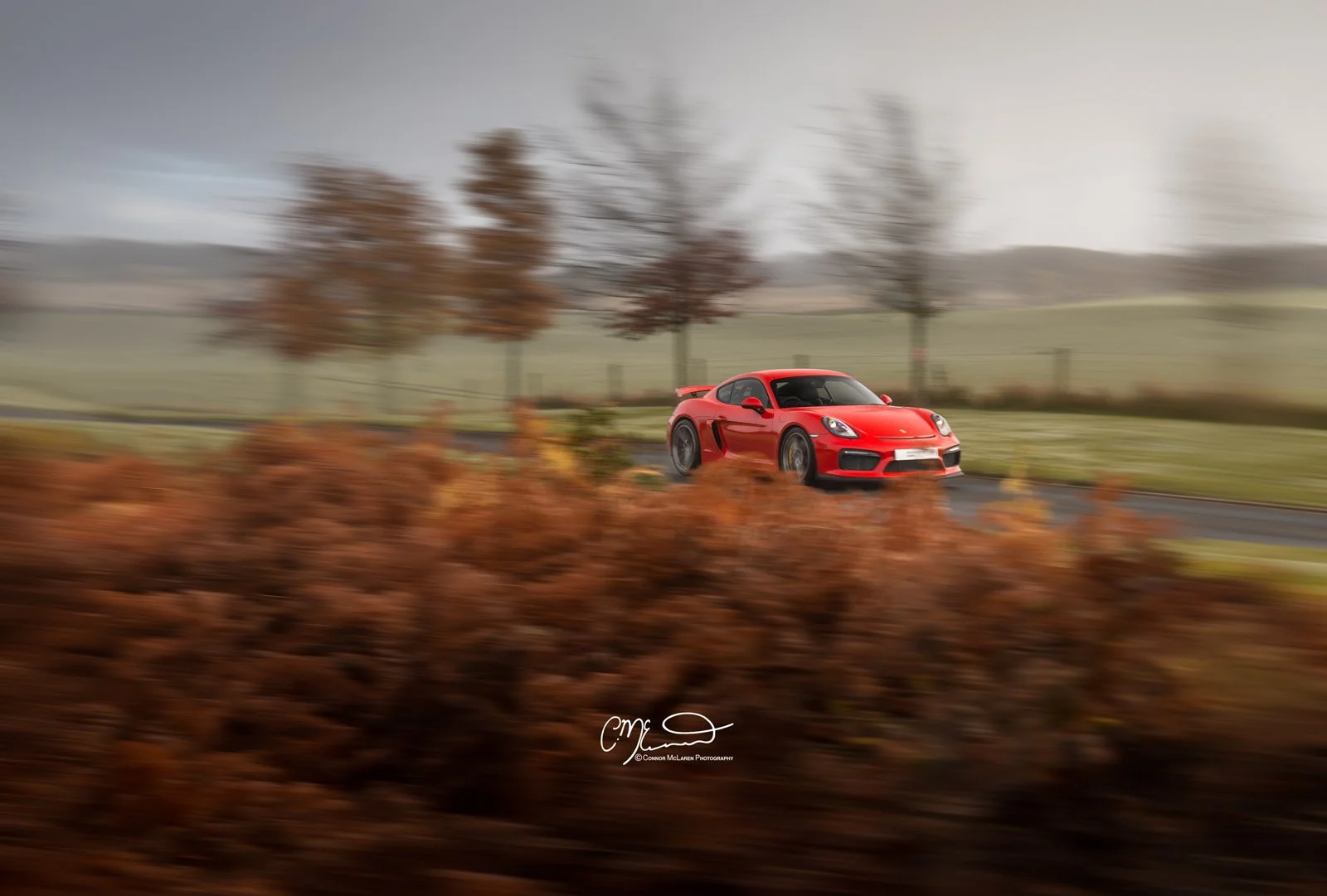 A red Porsche GT4 sports car driving on a blurred countryside road with trees and autumn foliage.