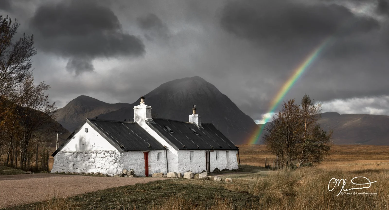 Blackrock cottage, glen coe, westeA white cottage with a black roof and chimneys in a rural landscape under a dark cloudy sky, with a rainbow extending from the ground to the clouds. Mountains are in the background, and trees are around the property.