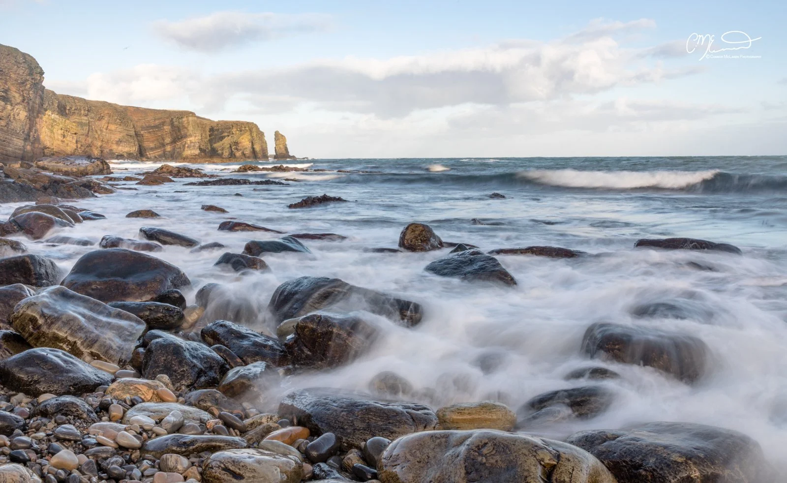 Orkney, Scotland. A rocky beach with waves washing over stones, cliffs in the background, and a partly cloudy sky.