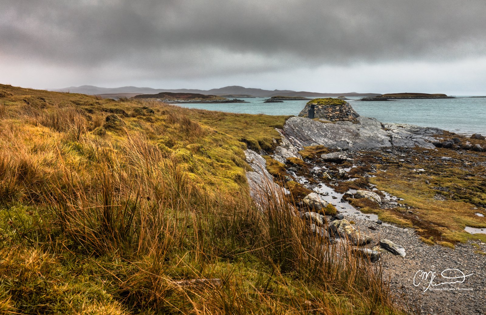 Scotland, Lewis Harris & Outer Hebrides, South Uist. Coastal landscape with grassy foreground, rocky shoreline, and small stone structure near the water under cloudy sky.