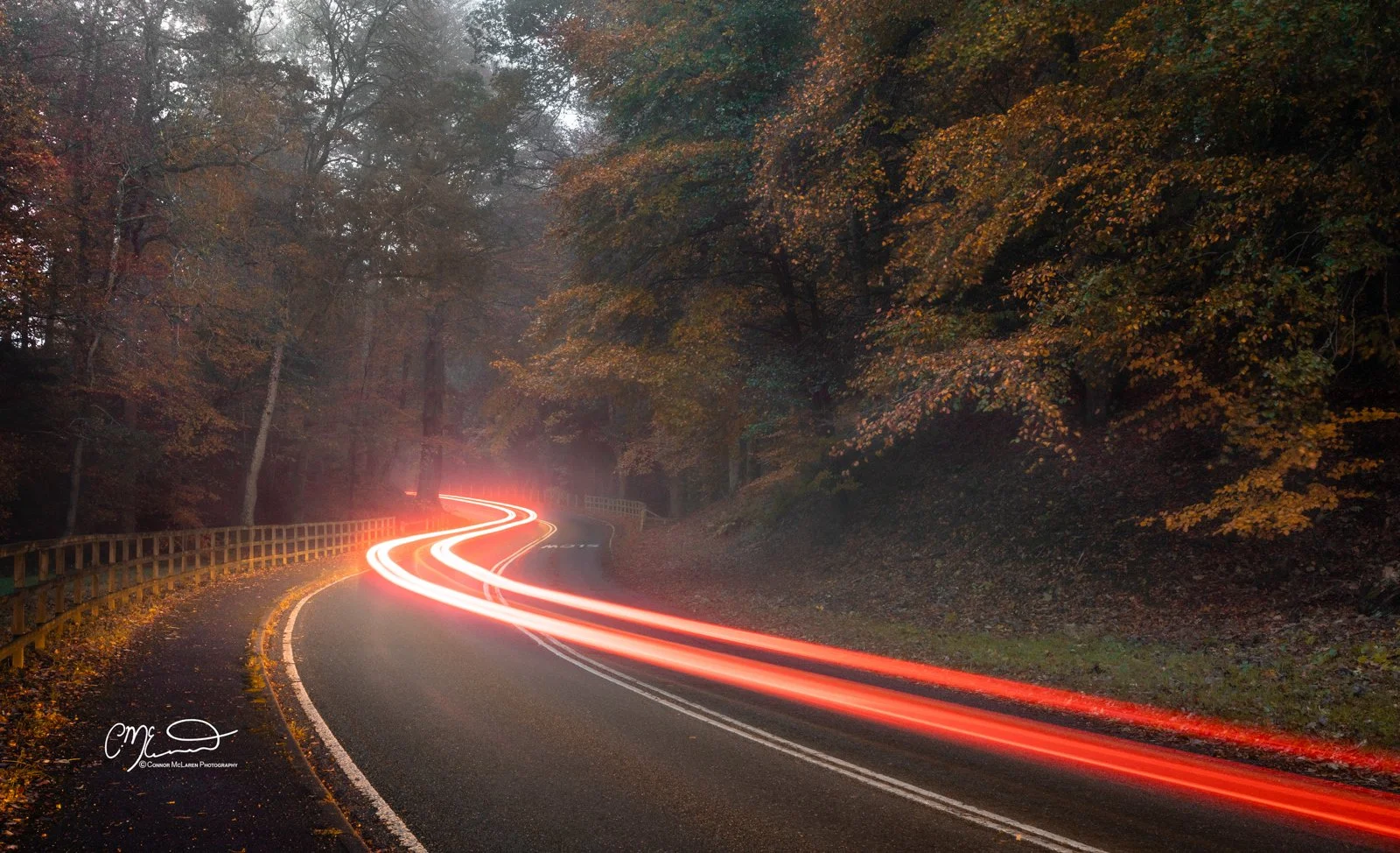 Scottish Central Highlands, Scotland, Perthshir. Curving mountain road through a forest with autumn-colored trees, light trails from moving vehicles, and foggy atmosphere.