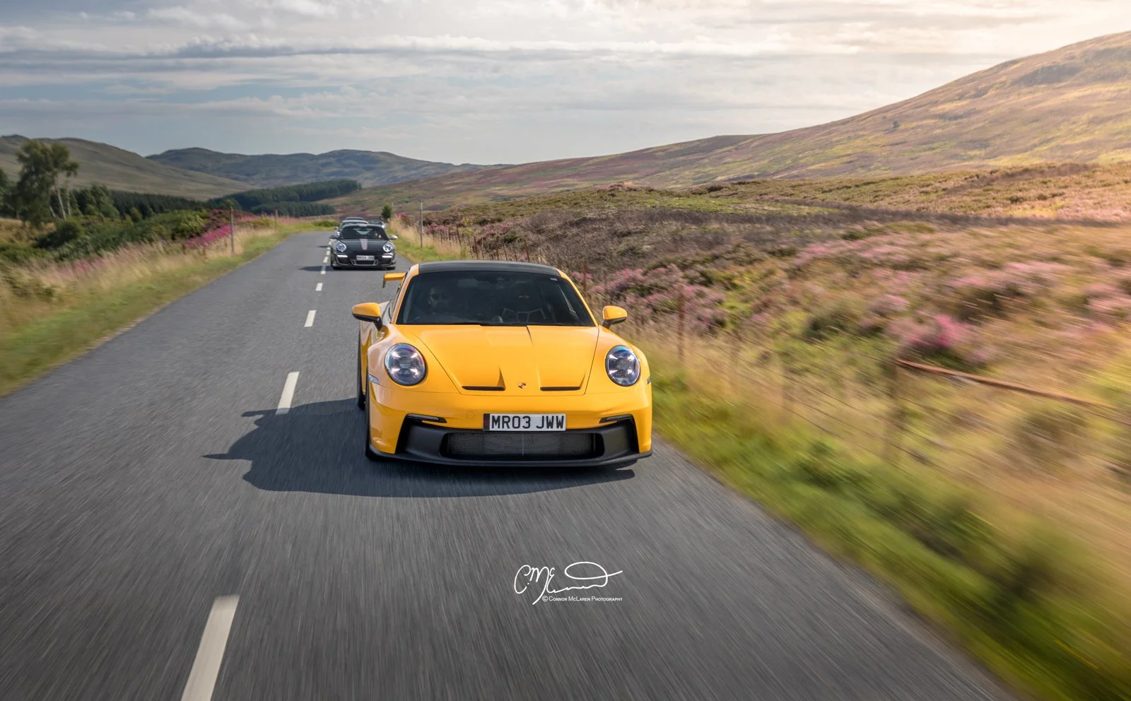 Mr JWW Porsche 911 GT3. Yellow sports car leading a line of several cars on a rural road through a scenic countryside with rolling hills and pink wildflowers.
