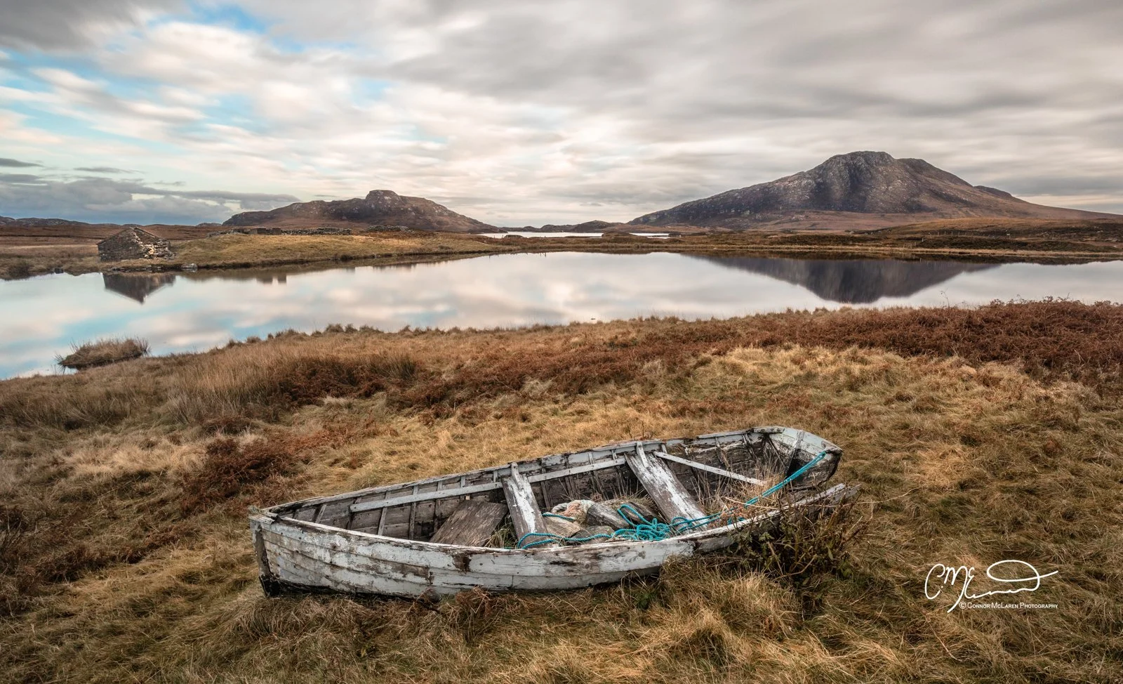 Scotland, Lewis Harris & Outer Hebrides, South Uist. An abandoned wooden boat on grassy terrain near a calm lake, with mountains and a cloudy sky in the background.
