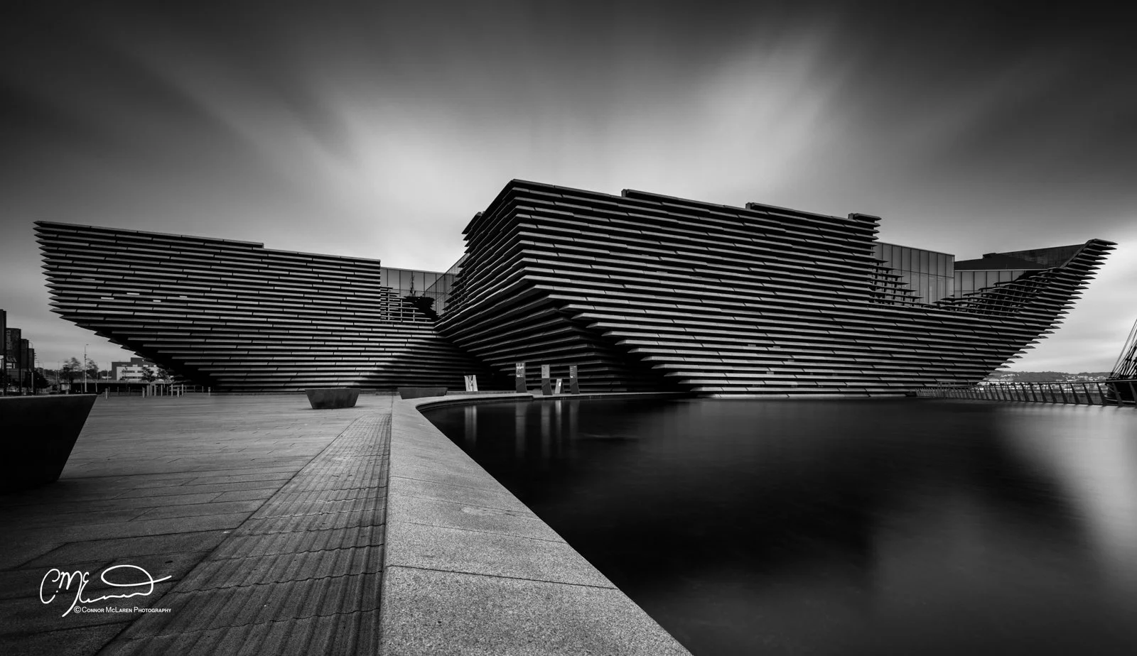 Scotland, Dundee, V&A. Black and white photo of a modern, angular building with layered horizontal slats, surrounded by a body of water, with a paved walkway in the foreground.