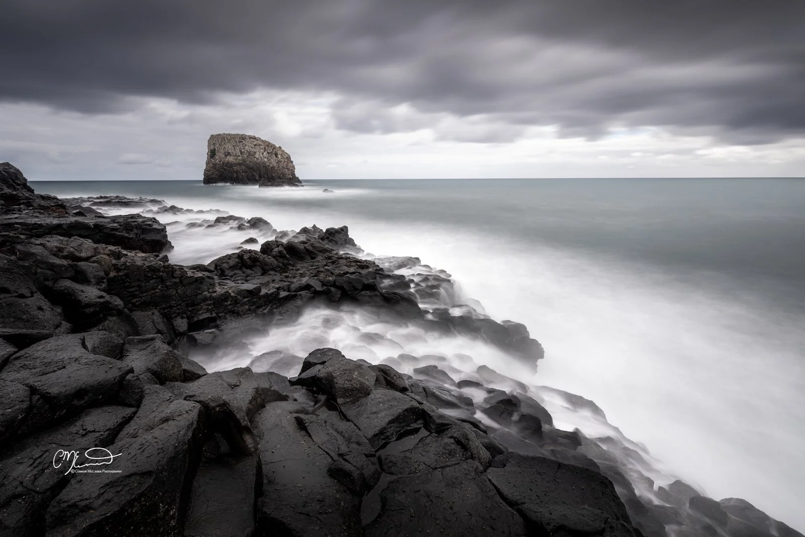 Volcanic rock, porto da cruz, Madeira Portugal. A rocky coastline with dark black rocks in the foreground, a large rock formation in the water, and stormy dark clouds overhead, creating a moody, overcast ocean scene.