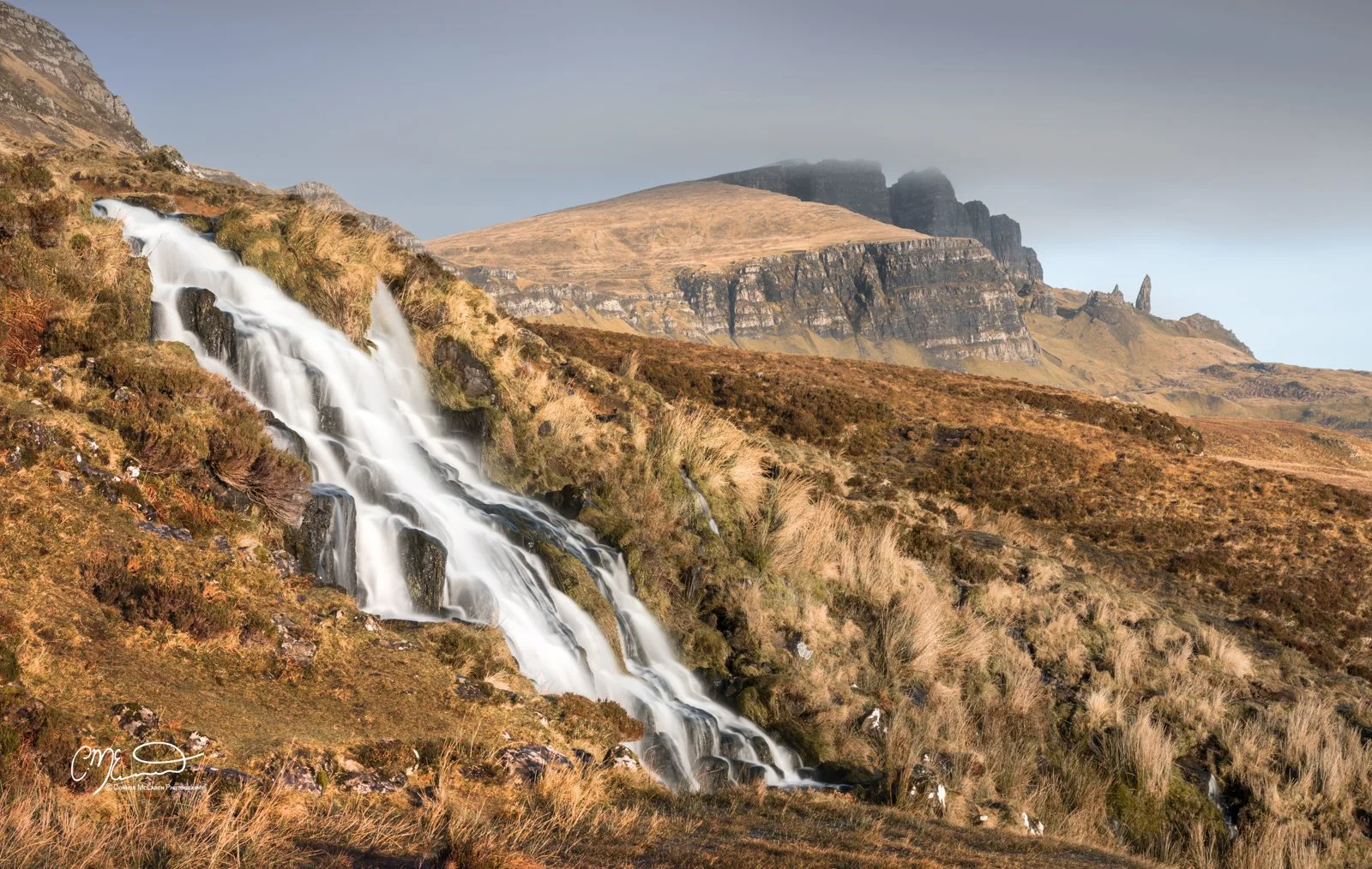 Scotland, Isle of Skye, Old Man of Storr. Scotland, Isle of Skye, Old Man of Storr. A waterfall flowing down a hillside with brown and green vegetation, with mountains in the background under a cloudy sky.